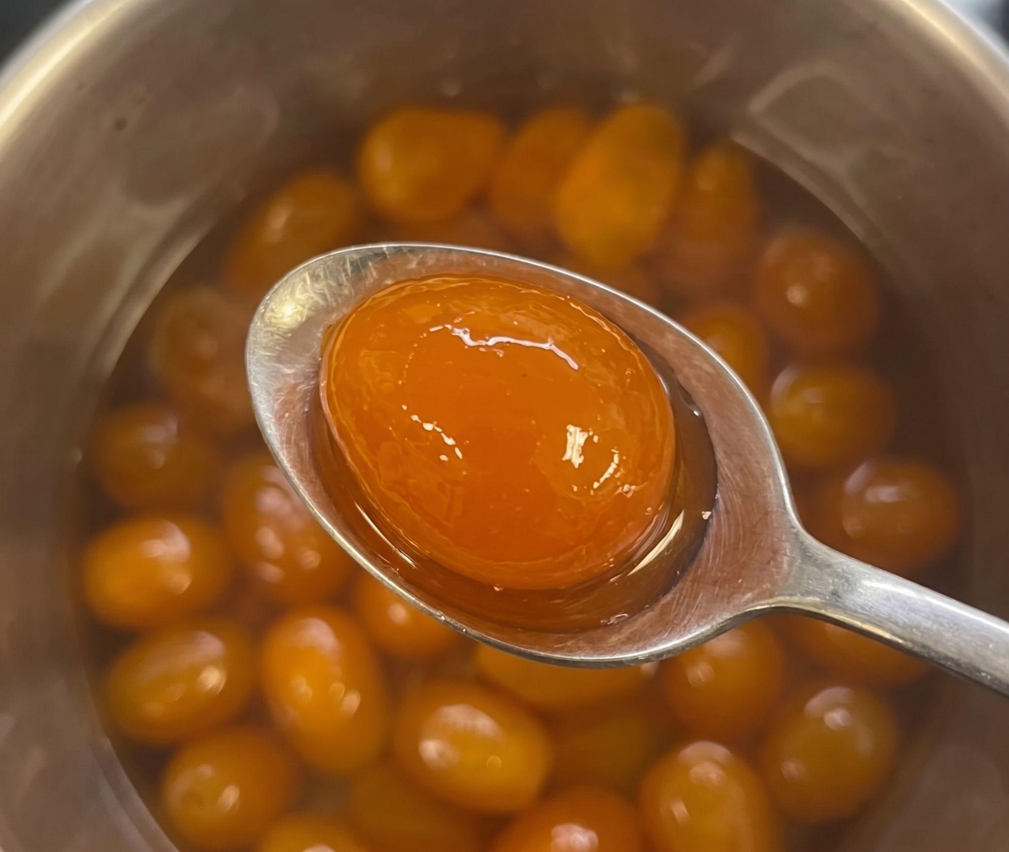 Spoon holding a glossy candied kumquat over a pot of confit kumquats